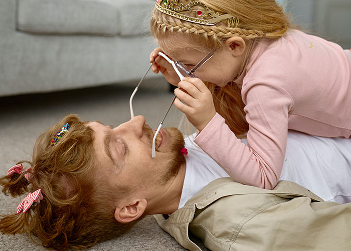 Padre acostado juguetonamente en el piso con su hijo, que lleva una tiara de la princesa y anteojos, mostrando el tiempo en familia.