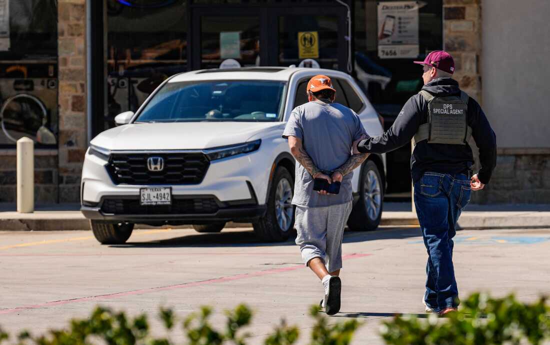 En esta foto, un agente federal y un hombre que está deteniendo son fotografiados desde atrás. El agente está en el lado derecho del marco, y está usando su mano izquierda para agarrar el brazo superior derecho del hombre detenido.