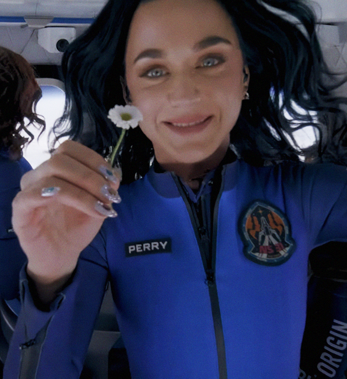 Mujer con traje azul sosteniendo una pequeña flor, sonriendo, con cabello oscuro; Parche visible en el atuendo.