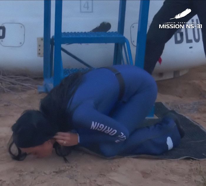 Persona con traje de espacio azul arrodillado en el suelo, con "Nuevo Shepard" y "Misión NS-31" visible, mostrando respeto.