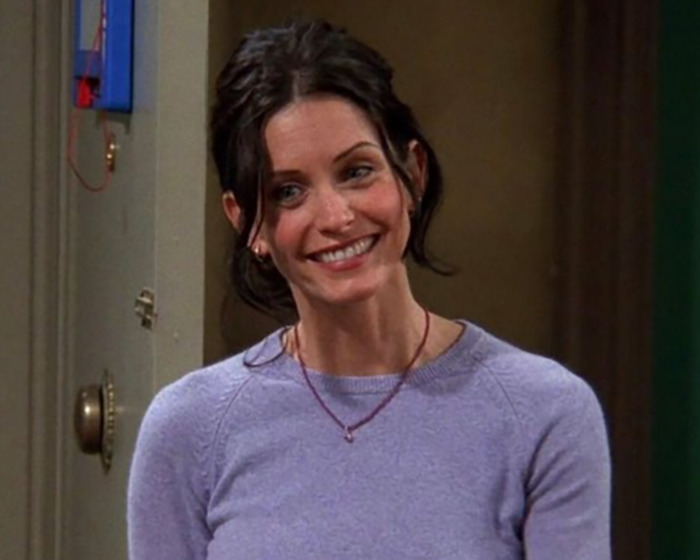 Ellen DeGeneres y Courteney Cox en el set durante un encuentro incómodo, sentado en sillas rojas con el logotipo del espectáculo en el fondo.