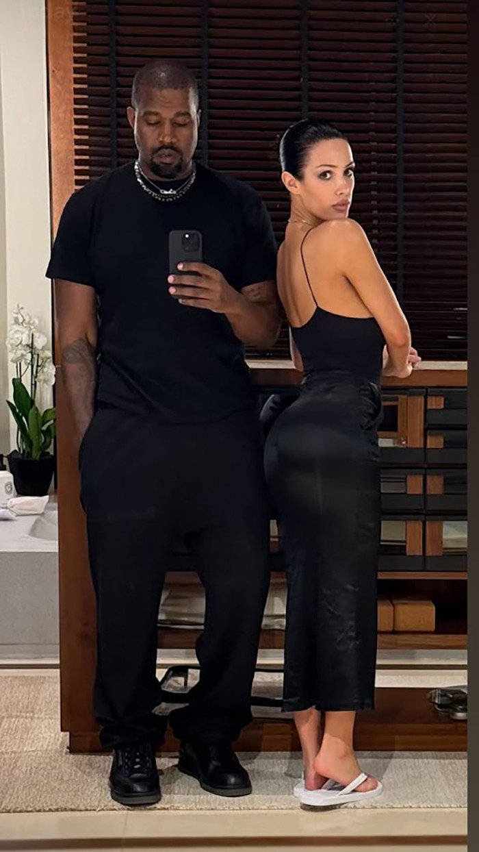 Una mujer de negro y un hombre de negro tomando una selfie espejo en casa, con persianas en el fondo.