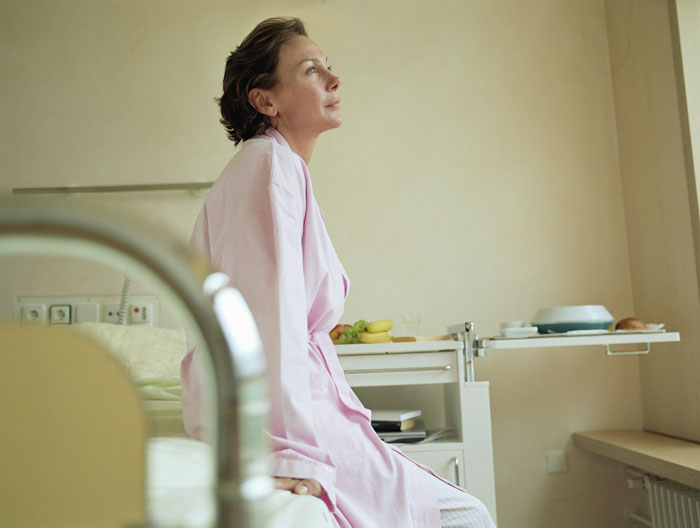 Mujer con una túnica rosa sentada en una cama de hospital, mirando cuidadosamente, reflexionando en una cena de compromiso.