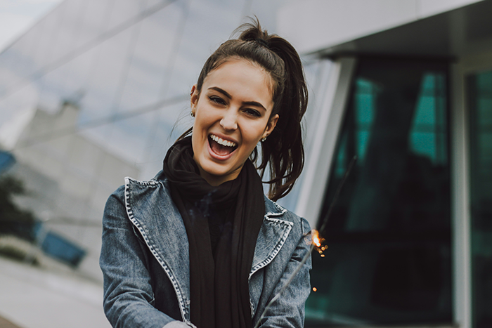 Mujer sonriendo intensamente frente a un edificio moderno, con una chaqueta de mezclilla y una bufanda negra.