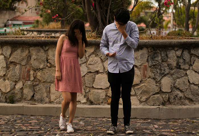 Una mujer y un hombre se separan, ambos cubriendo sus caras, en un parque con muro de piedra y flores.