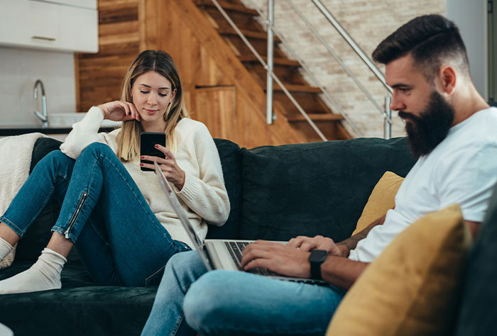 Pareja sentada en un sofá, mujer mirando su teléfono, hombre usando una computadora portátil, discutiendo la frustración de la planificación de la boda.