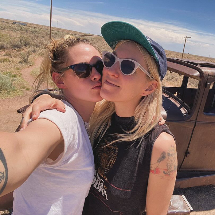Los tortolitos comparten un beso al aire libre, con gafas de sol y vestimenta informal, posando junto a un automóvil antiguo.