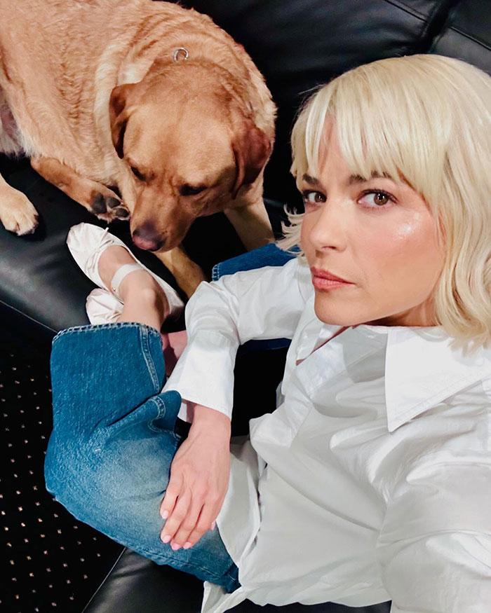 Selma Blair muestra su personalidad en una camisa blanca, sentada en un sofá con un perro, exudando encanto y confianza.