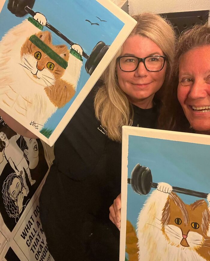 Dos personas sonriendo con pinturas de un gato que anteriormente levantaba pesas.