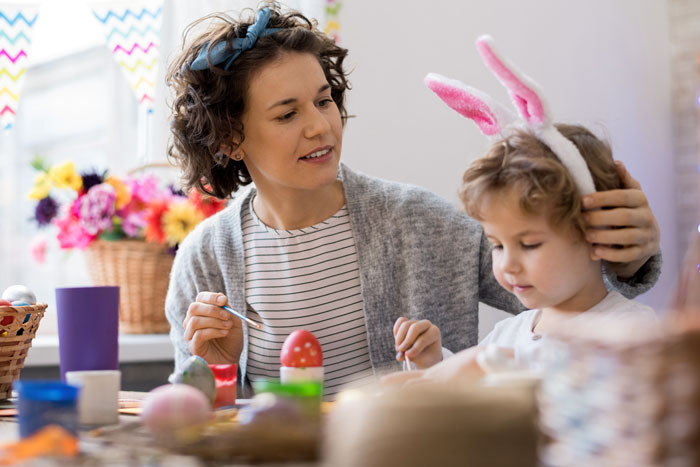 Mujer e hijos con orejas de conejo pintando huevos de Pascua en un entorno brillante y festivo.