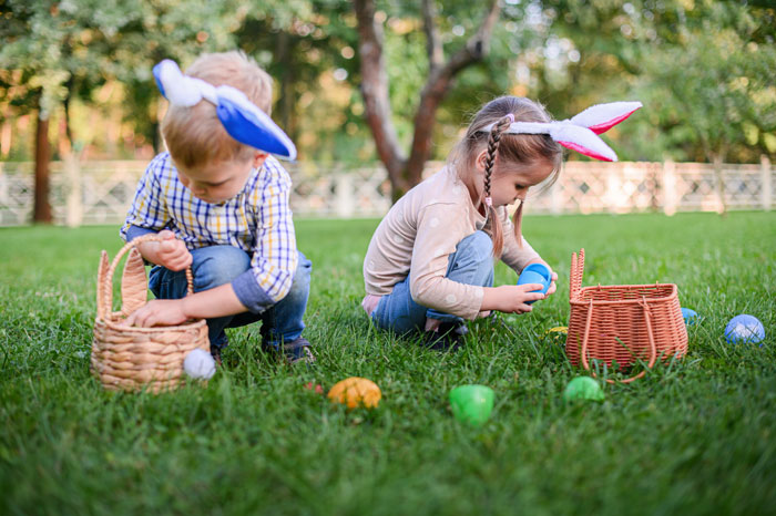 Dos niños con orejas de conejito en un campo cubierto de hierba recolectando huevos de Pascua, destacando una alegría de Pascua.