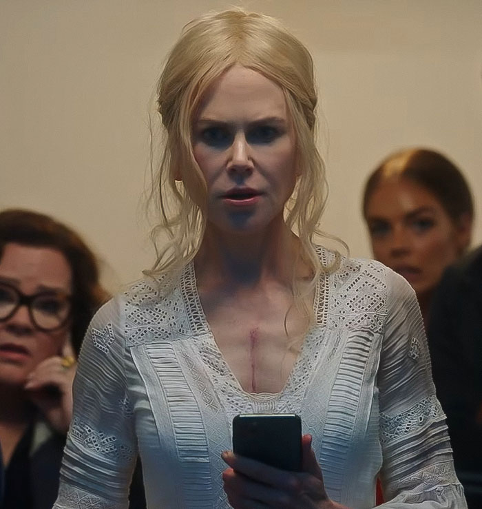 Nicole Kidman, de 57 años, con un top de encaje blanco, sosteniendo un teléfono, con personas preocupadas en el fondo.