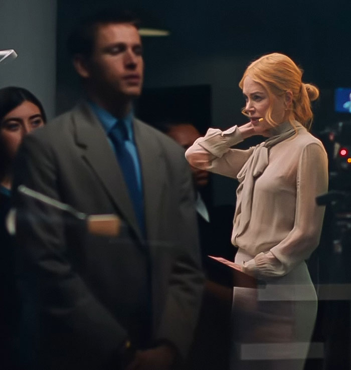 Nicole Kidman, de 57 años, en una escena seria con blusa ligera y falda, capturada en una habitación con poca luz con figuras borrosas.