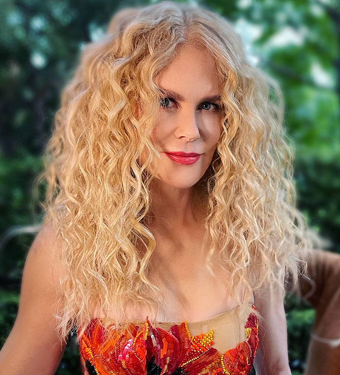 Nicole Kidman con cabello rubio rizado y lápiz labial rojo posando al aire libre con colorido vestido floral.