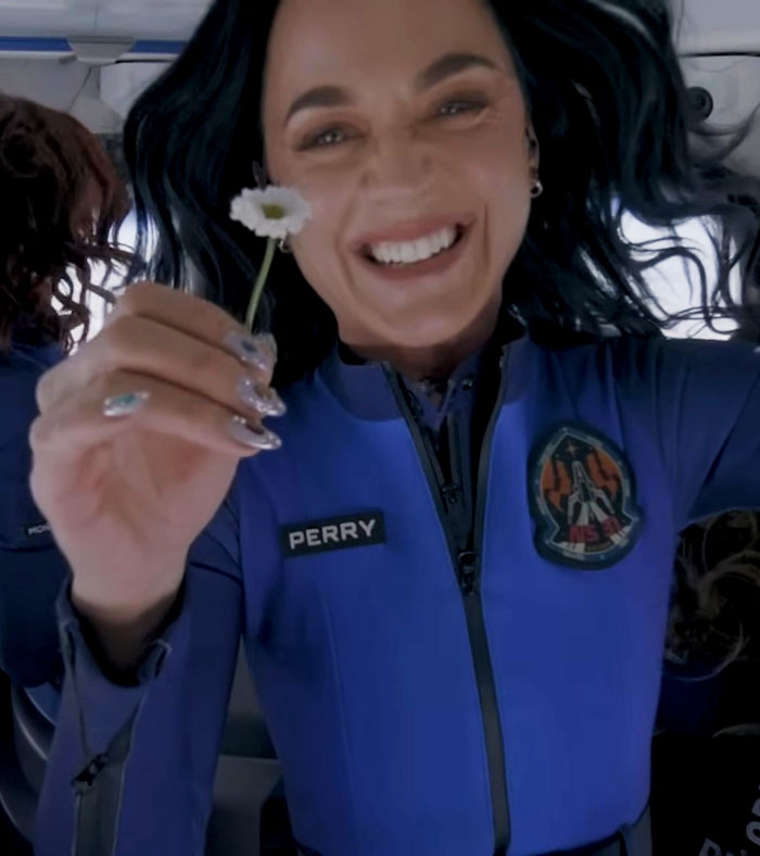 Katy Perry sonriendo con traje de vuelo de origen azul que sostiene una pequeña flor durante la experiencia de vuelo espacial.