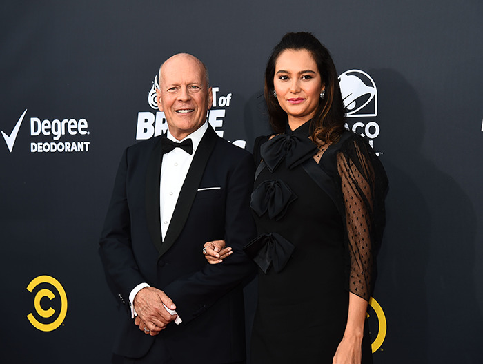 Bruce Willis con su esposa en una alfombra roja con atuendo formal, sonriendo para una foto juntos.