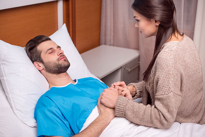 Hombre en la cama de ER mientras su novia sostiene su mano, teléfono cerca, tensión en el aire.