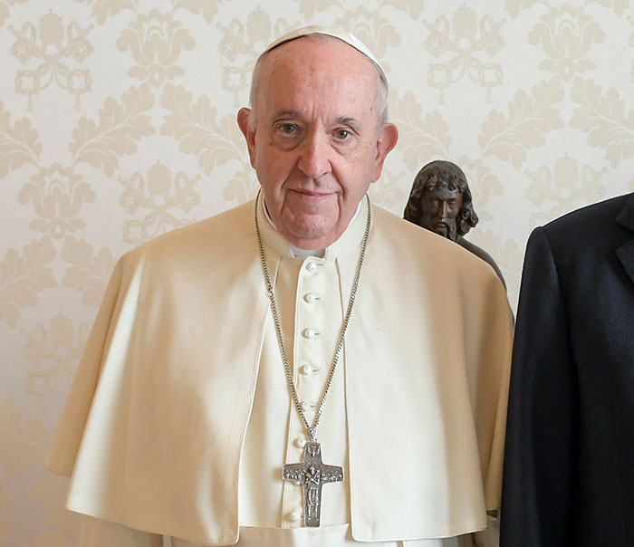 El Papa Francisco en una bata blanca, de pie en el interior con una expresión serena, capturando reacciones mixtas.