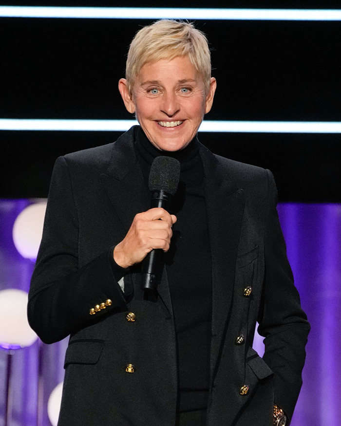 Ellen DeGeneres sosteniendo un micrófono, con un traje negro, sonriendo en el escenario.