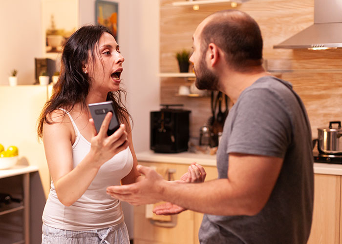 Mujer sosteniendo el teléfono discutiendo con el esposo en la cocina por el horario de los niños.