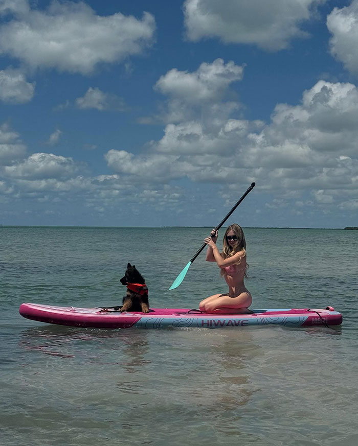 Sydney Sweeney en Paddleboard con cachorro en arnés rojo, en agua tranquila bajo un cielo parcialmente nublado, disfrutando de actividades al aire libre.