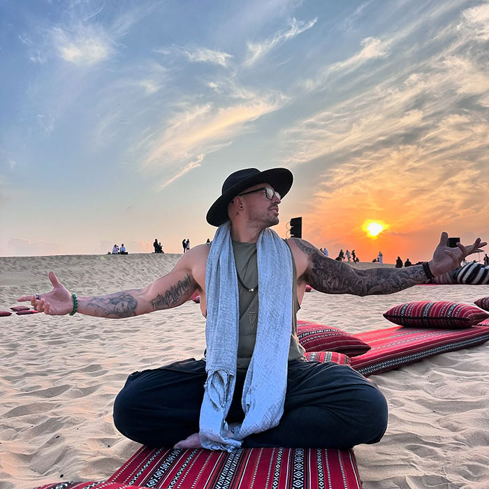 Hombre con tatuajes y sombrero meditando al atardecer en la arena del desierto, lo que refleja la ex bailarina del pole convertida en la historia de los monjes.