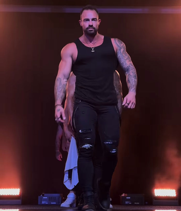 Hombre musculoso con tatuajes y camiseta sin mangas negra caminando en el escenario, simbolizando a la ex bailarina del pole convertida en una historia de monje.