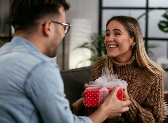 La mujer sorprende al marido con la prueba de embarazo como regalo de cumpleaños, ambos comparten un momento emocional en un acogedor sofá en el interior.