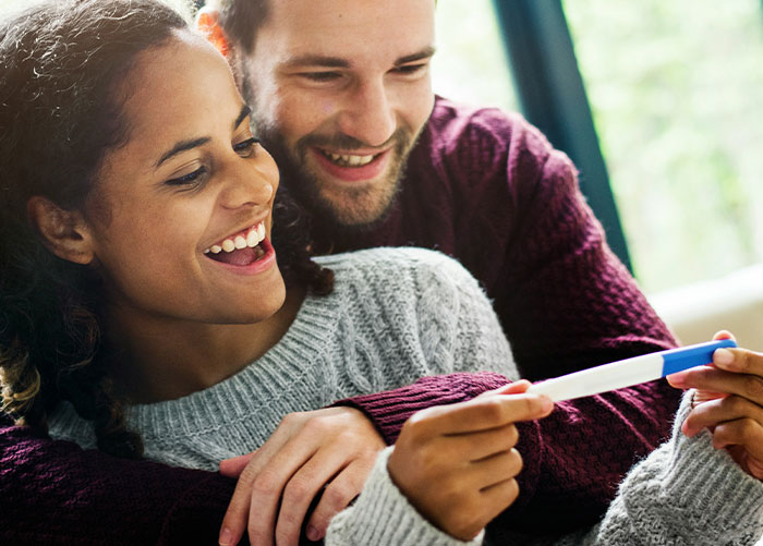 La mujer sorprende al marido con la prueba de embarazo como regalo, sonriendo y compartiendo un momento alegre en casa.