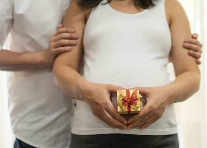 Mujer embarazada sosteniendo una pequeña caja de regalo con su esposo parado, simbolizando el regalo sorpresa de la prueba de embarazo.
