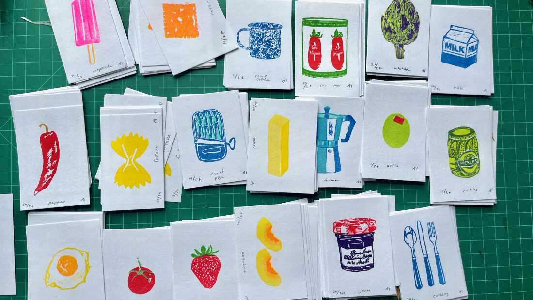 Tres hileras de impresiones hechas por la artista Ana Inciardi muestran artículos coloridos como una taza, estaño de sardina, paleta, alcachofa, pasta de espino, chile rojo, oliva, un frasco de mermelada, duraznos, una fresa y un huevo frito.