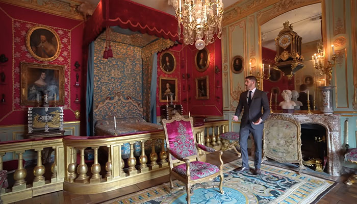 El hombre en traje dentro de la casa más cara del mundo con muebles adornados, decoración roja y dorada, y pinturas clásicas en las paredes.