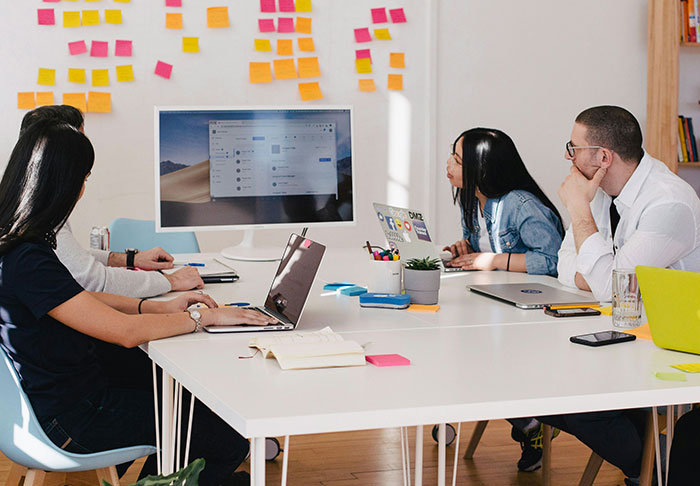 Cuatro compañeros de trabajo en una oficina moderna con computadoras portátiles y notas adhesivas, discutiendo una nominación de trabajo arriesgada sin hijos.