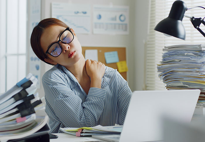 Mujer cansada en la oficina frotando su hombro rodeada de papeleo, ilustrando una situación laboral extra riesgosa sin hijos.