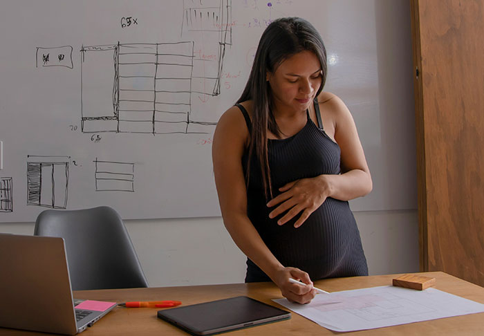 Mujer embarazada que trabaja en un proyecto en el escritorio con computadora portátil y pizarra, ilustrando tareas de trabajo extra riesgosas sin hijos.