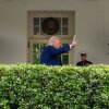 En esta foto, el presidente Trump saluda con una mano mientras está fuera de la Casa Blanca. Una fila de setos está en primer plano.