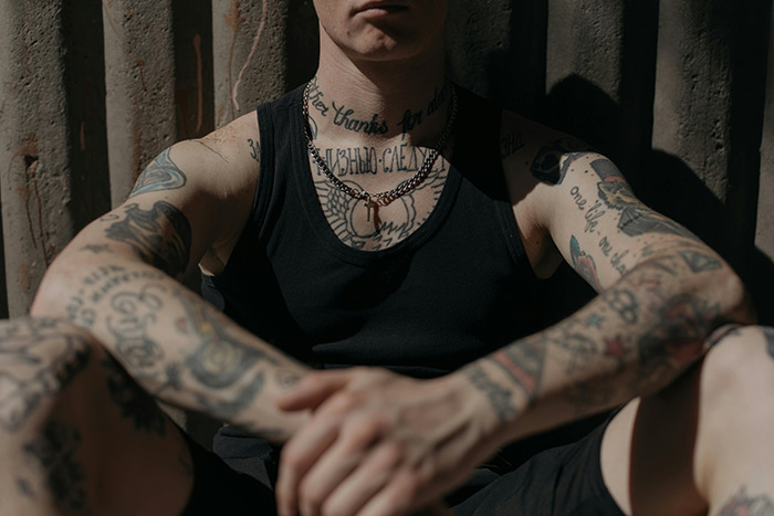 Persona con múltiples tatuajes en los brazos y el cuello, sentado contra un fondo de textura oscura a la luz del sol suave.