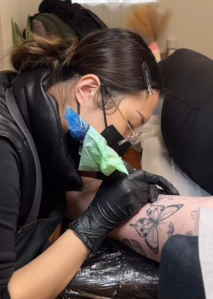 Artista del tatuaje con guantes y máscara creando un tatuaje detallado de mariposas en el brazo de un cliente durante el boom del tatuaje.