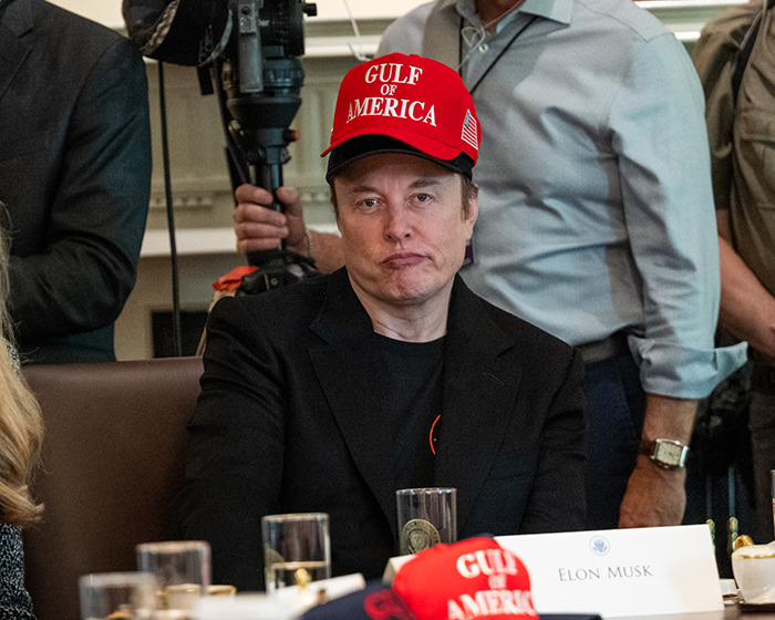 Elon Musk con un sombrero rojo del Golfo de América, sentado en una mesa de reuniones con una expresión seria.