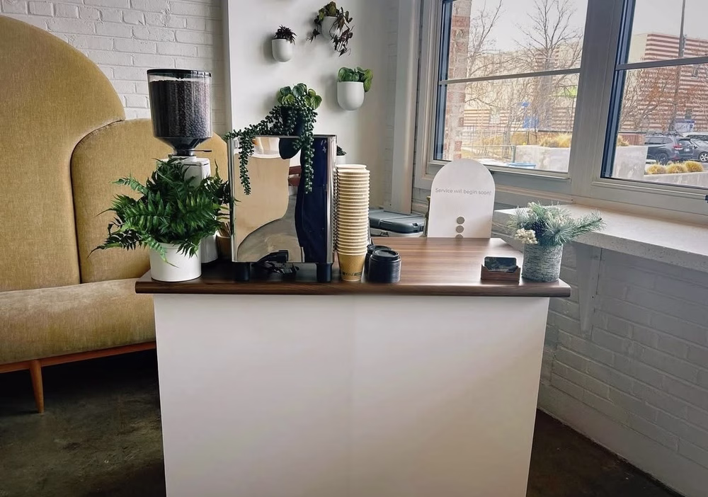 Un carrito de café de Goodhart Coffee está cubierto con plantas, una pequeña máquina de café espresso, un molinillo de café y tazas de café y tapas para llevar.