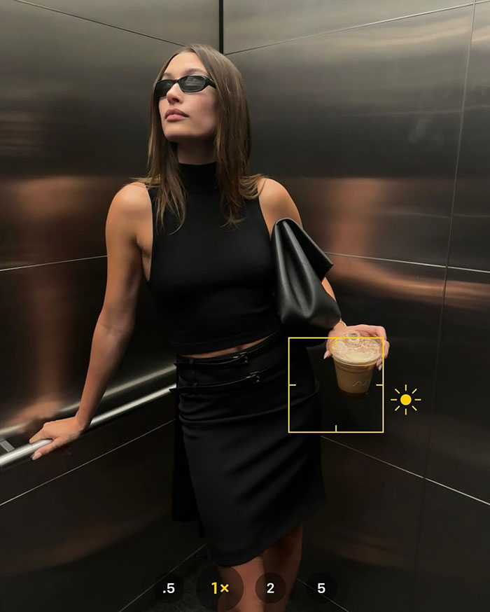 Hailey Bieber con atuendo negro y gafas de sol posando en ascensor sosteniendo una bebida después del berrinche del juego de hockey de su esposo Justin