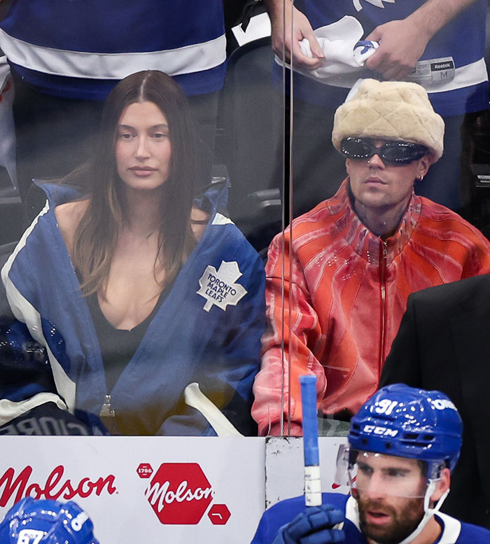 Hailey Bieber mirando el juego de hockey con chaqueta azul al lado de Justin con gafas de sol y sombrero de piel en la arena.