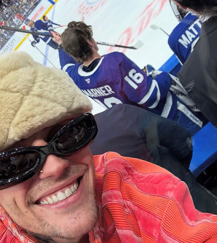 Justin Bieber sonriendo en un juego de hockey, con gafas de sol y un sombrero peludo, con jugadores en el hielo en el fondo.