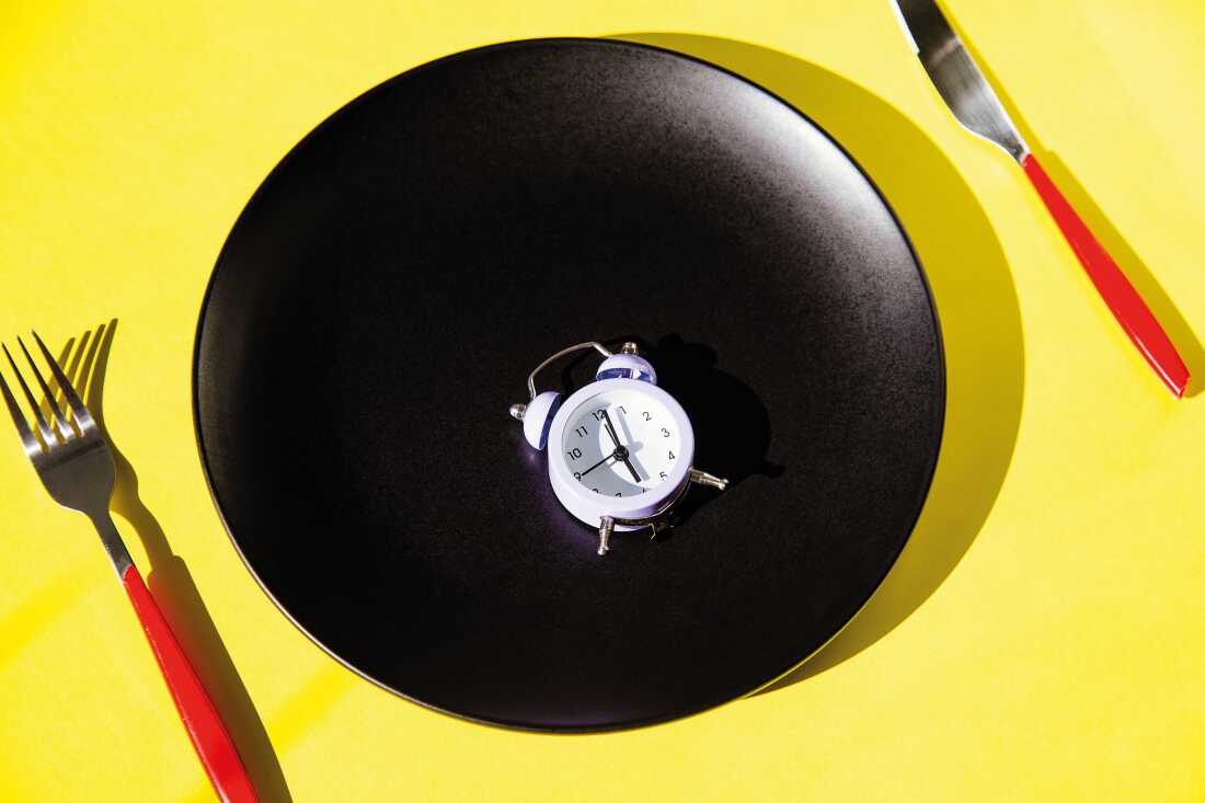 La fotografía de estudio muestra un plato de cerámica grande y negro sobre un fondo amarillo. Una horquilla de metal y un cuchillo con manijas de plástico rojo están a la derecha e izquierda de la placa y AA Small White Simut Watlock descansa sobre la placa negra.