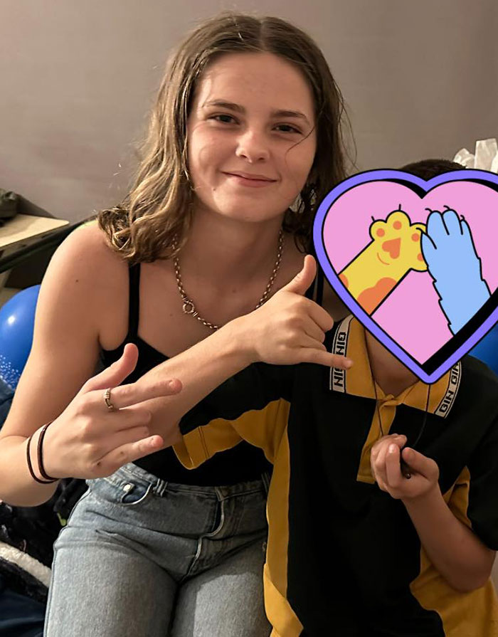 La adolescente sonriente haciendo gestos con las manos mientras está sentada al lado de un niño con un emoji del corazón que se cubre la cara.