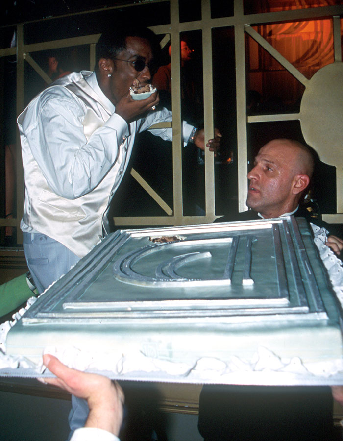 El hombre comiendo pastel desordenadamente mientras otra persona sostiene el pastel grande, destacando los repugnantes hábitos alimenticios de Diddy revelados por el personal.