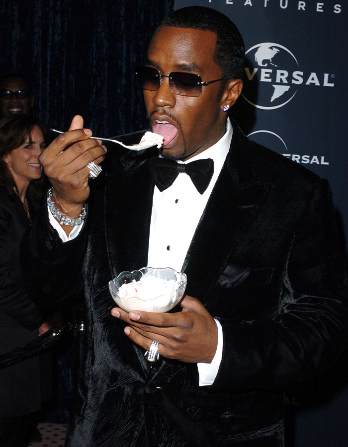 Diddy con gafas de sol y un esmoquin negro, comiendo postre de un tazón de vidrio en un evento formal.