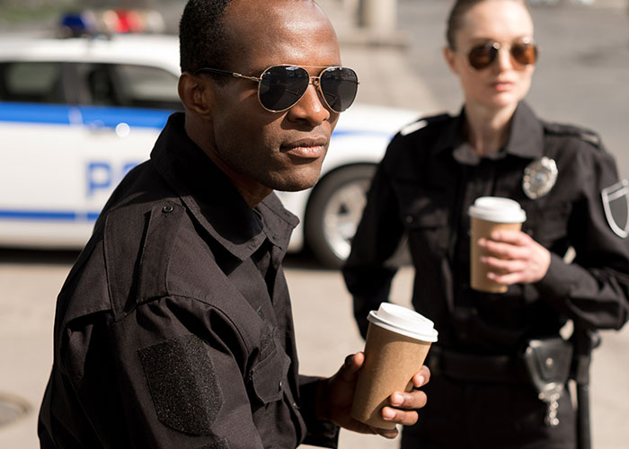 Dos policías que usan gafas de sol con tazas de café para llevar afuera cerca de un vehículo policial.