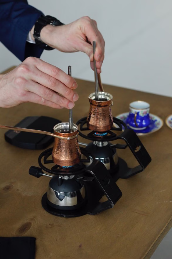 Un primer plano de un café barista que elabora café en dos cezves de cobre, que se calientan por una llama baja en dos quemadores.