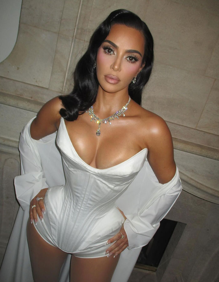 Kim Kardashian posando en un corsé blanco y joyas en un entorno glamoroso, con los fanáticos discutiendo sus cirugías de plástico.